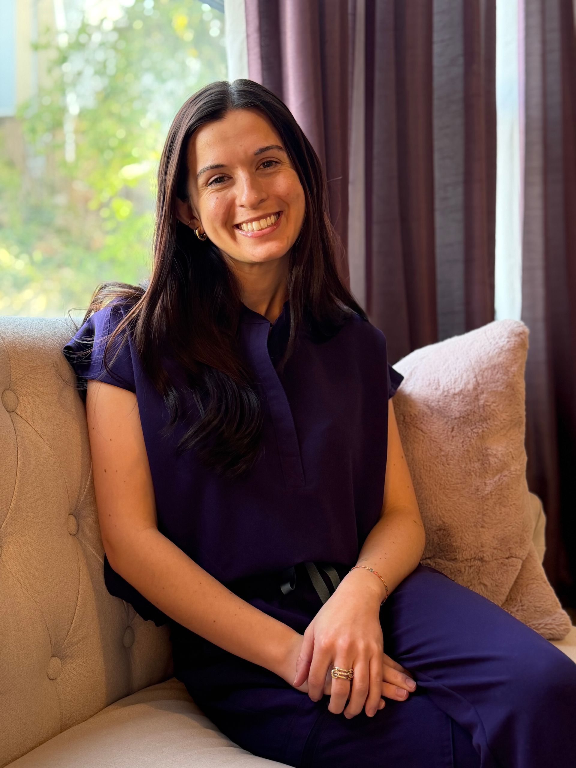 Smiling woman in a purple outfit sitting on a beige couch by a window with curtains.