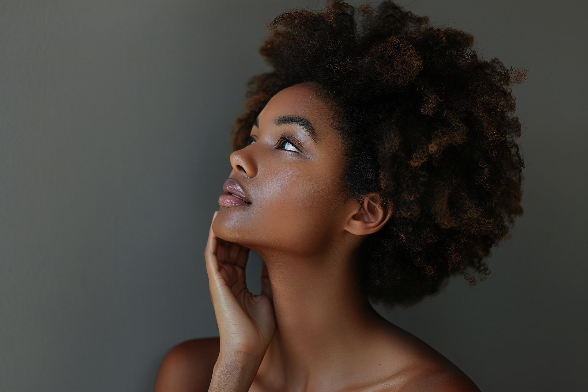 Portrait of a person with curly hair looking upward, hand resting on chin, against a gray background