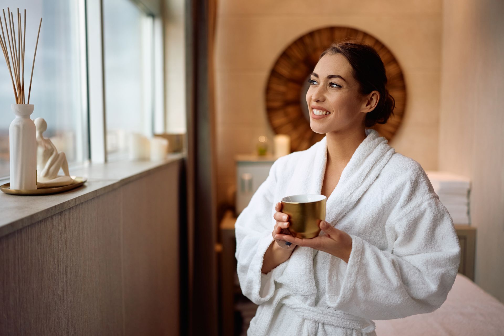 Woman in a white robe holding a cup in a spa room by a window, smiling softly