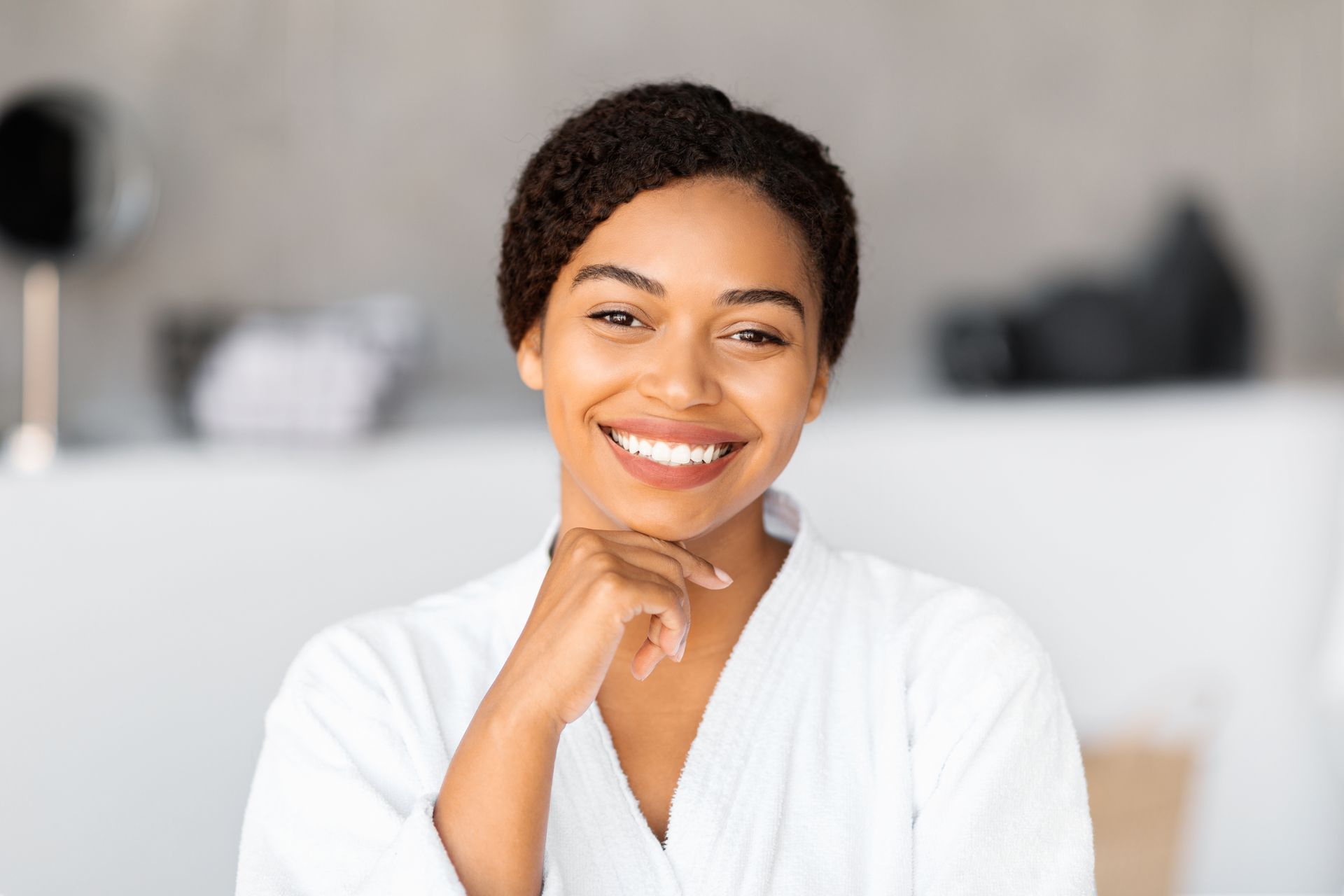 Smiling person in a white robe resting chin on hand, seated in a bright room with a blurred background