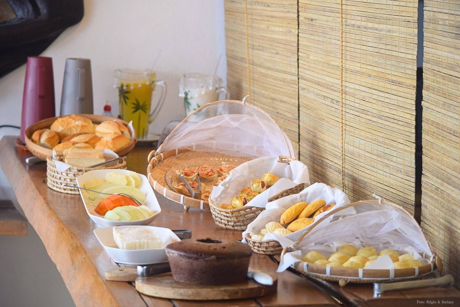 Buffet de café da manhã com diversos pães, doces e queijos dispostos em uma mesa de madeira.
