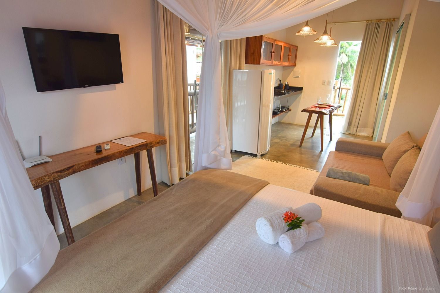 Quarto com cama, sofá, cozinha compacta e acesso à varanda. Tons de branco e bege.
