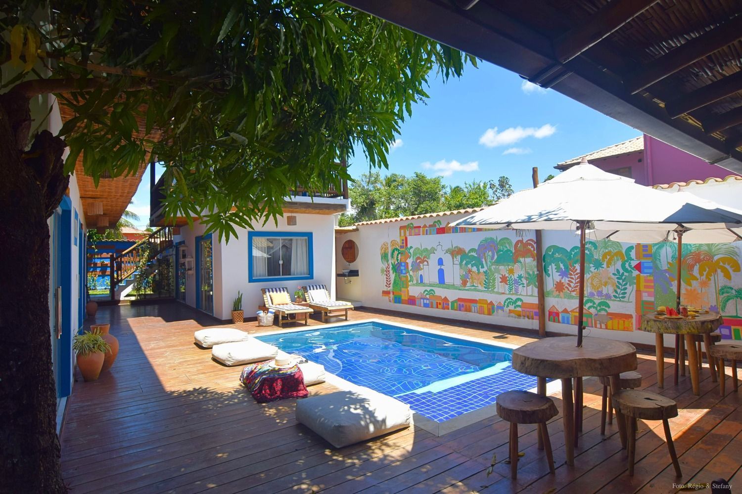 Pátio com piscina, assentos, mural e deck de madeira. Dia ensolarado.