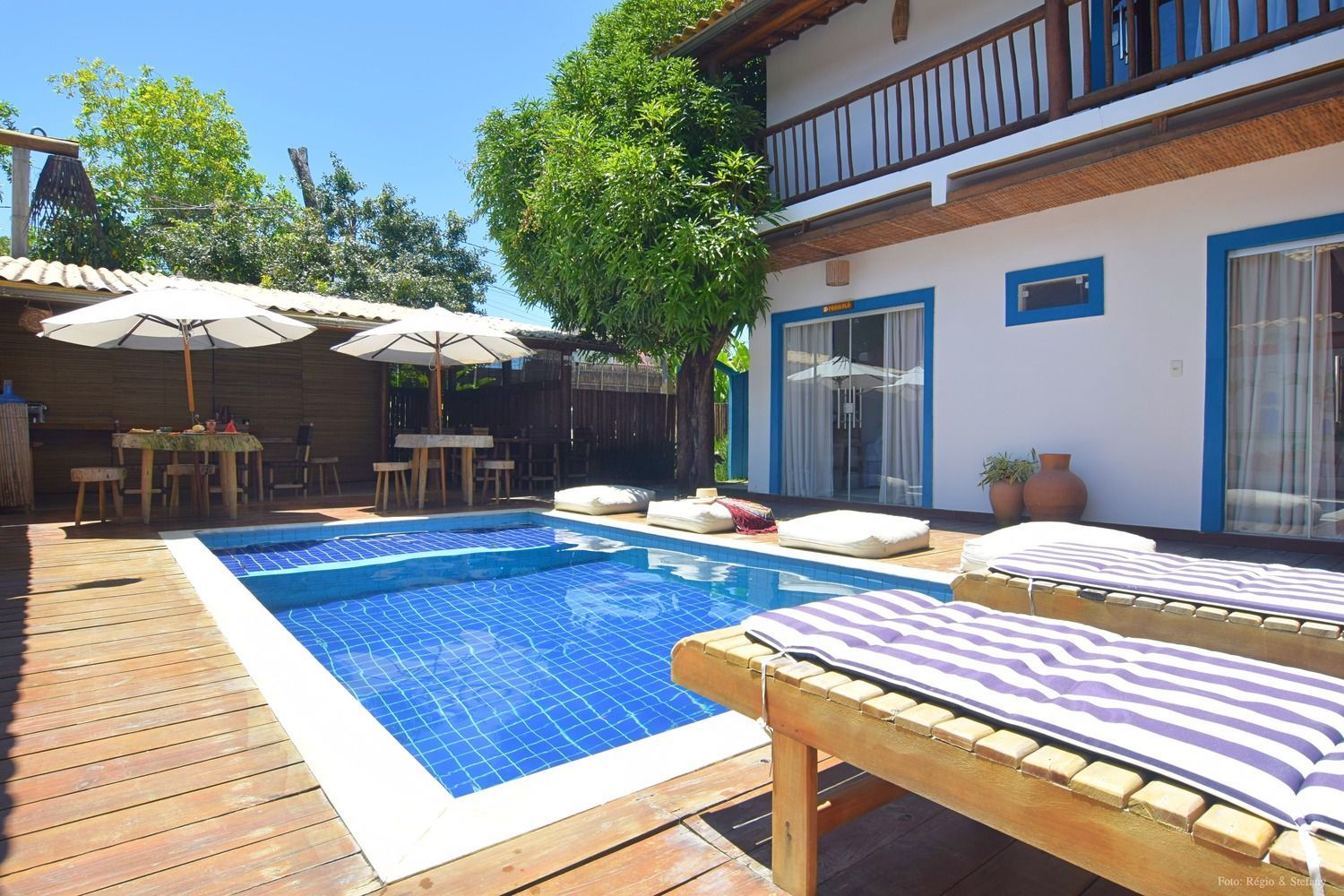 Vista da piscina com um prédio branco com detalhes em azul, deck de madeira, espreguiçadeiras e guarda-sóis sobre as mesas.