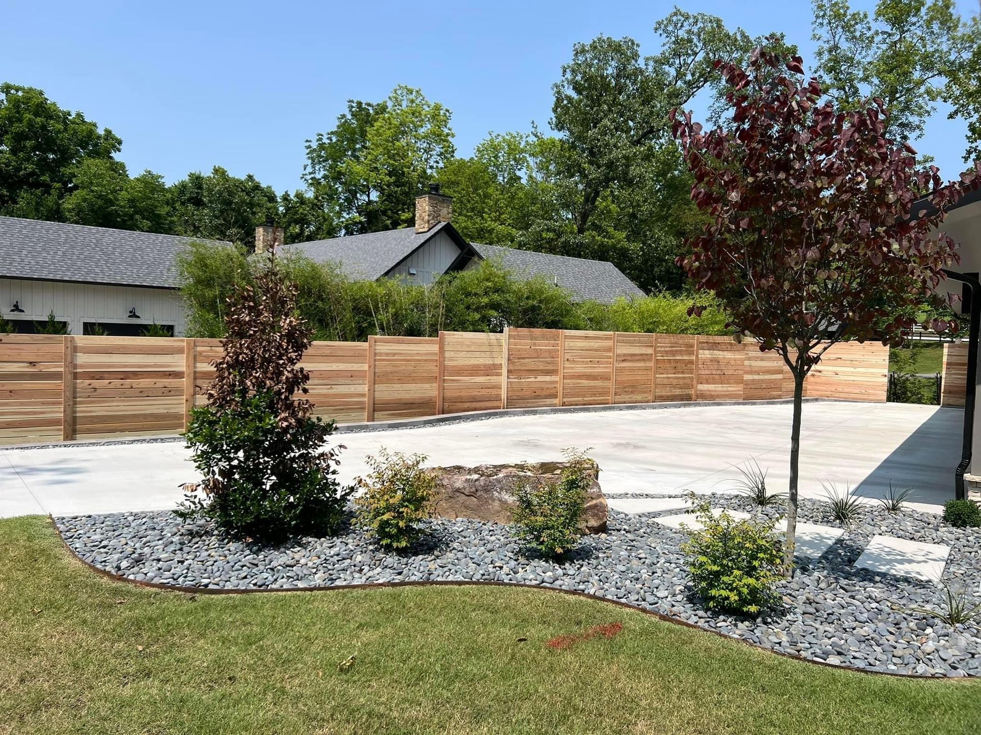 Landscaped front yard with small trees, shrubs, gravel beds, and a wooden fence by a house
