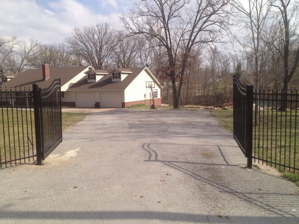 Open black gates leading to a paved driveway toward a small house with bare trees behind it.