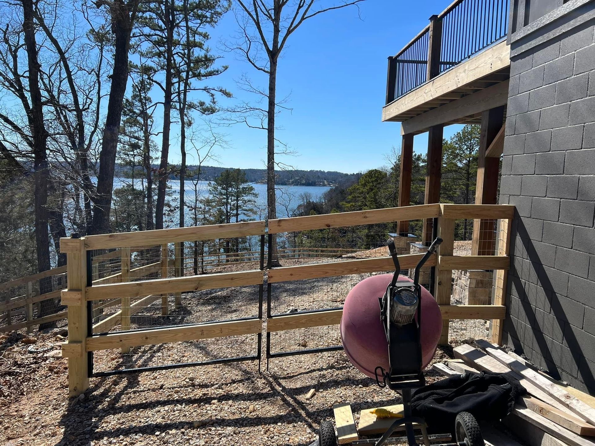 Wooden deck railing overlooking a lake, with stacked boards and tools beside a gray house under a clear blue sky
