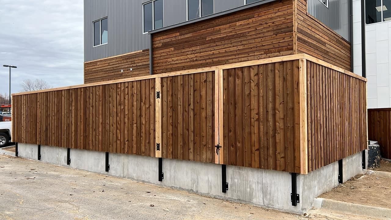Wooden privacy fence around a gray building on a concrete foundation by a gravel lot
