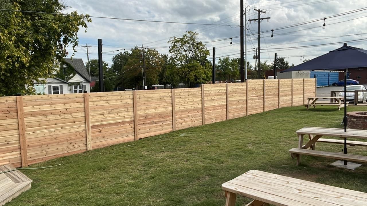 Wooden privacy fence lining a grassy park area with picnic tables and trees under a cloudy sky