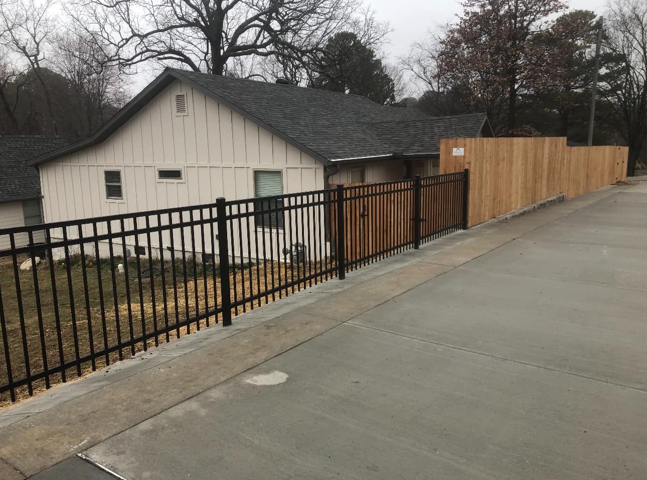 Concrete path beside black metal fence and beige building with wooden privacy fence under overcast sky