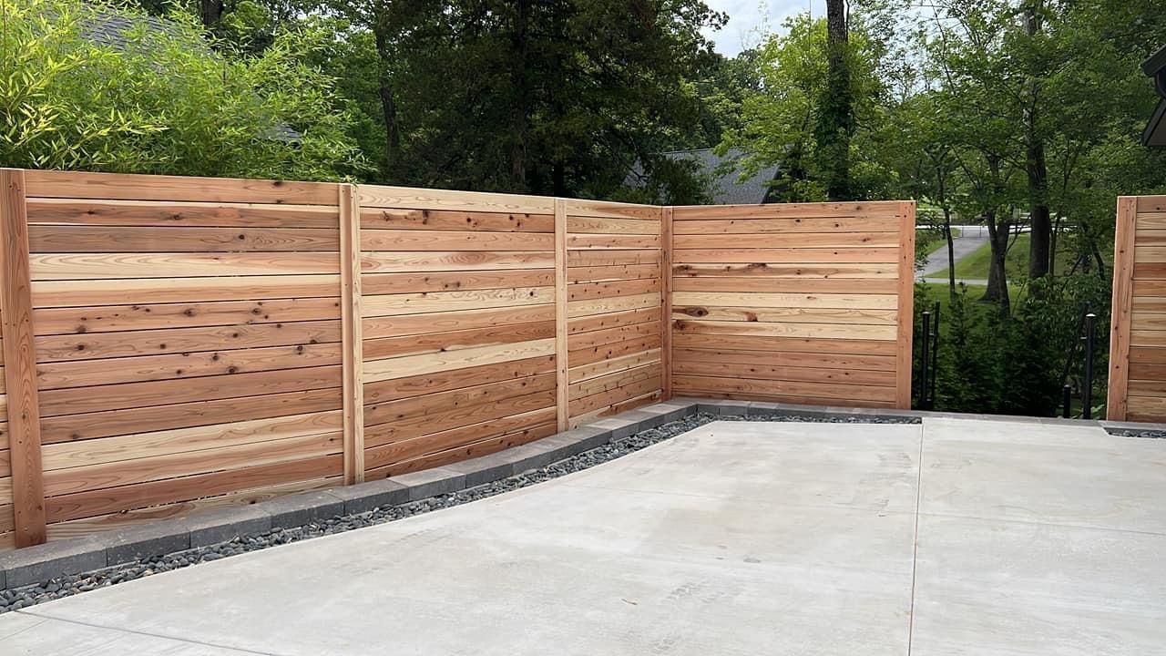 Wooden privacy fence along a concrete driveway with trees in the background