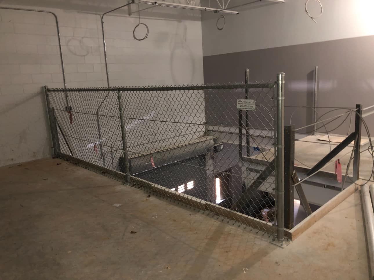 Chain-link fenced industrial pit with stairs and exposed pipes in a concrete basement
