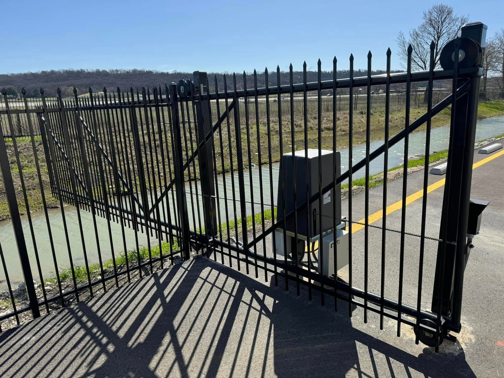 Black metal gate across a paved path, with shadows cast in bright sunlight.