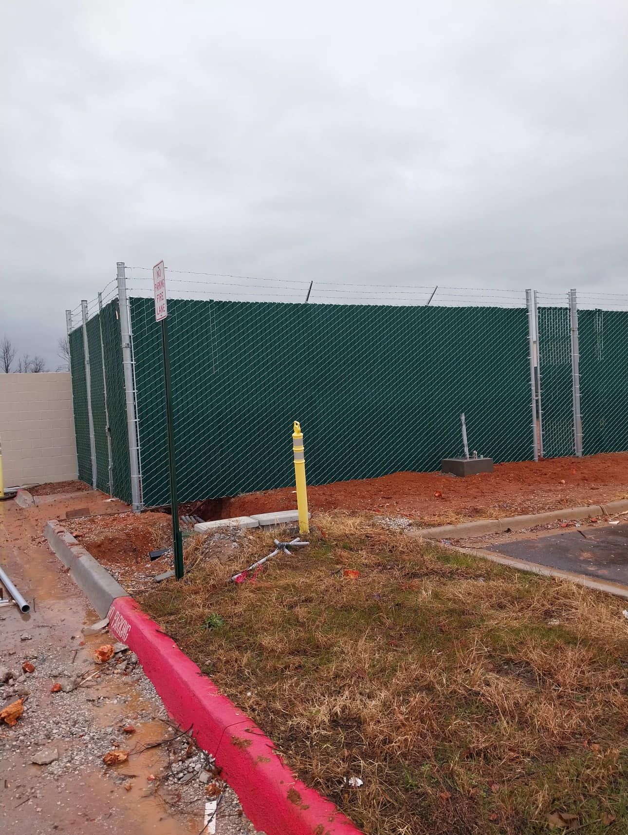Green fenced enclosure by a roadside, bordered by a red curb and wet pavement in a cloudy setting