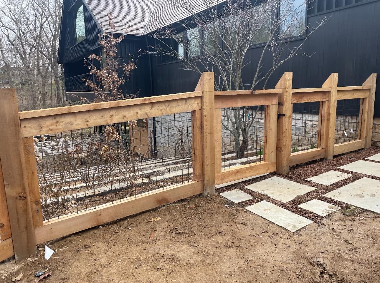Wooden fence with wire mesh beside a dirt yard and stone pavers, in front of a house and trees