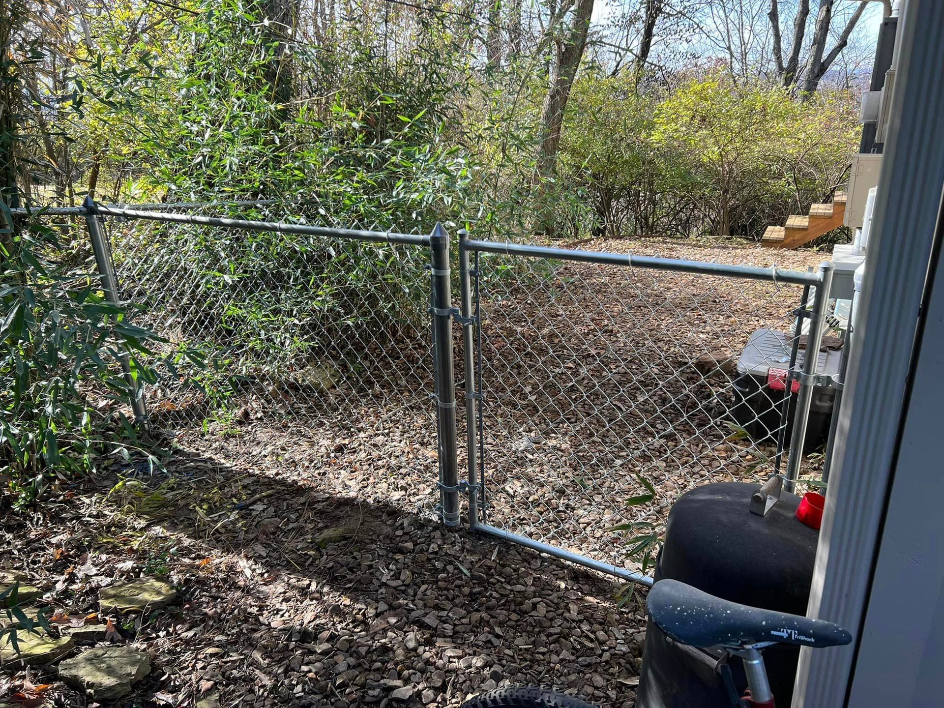 Chain-link gate opening to a wooded backyard with a black grill in the foreground.