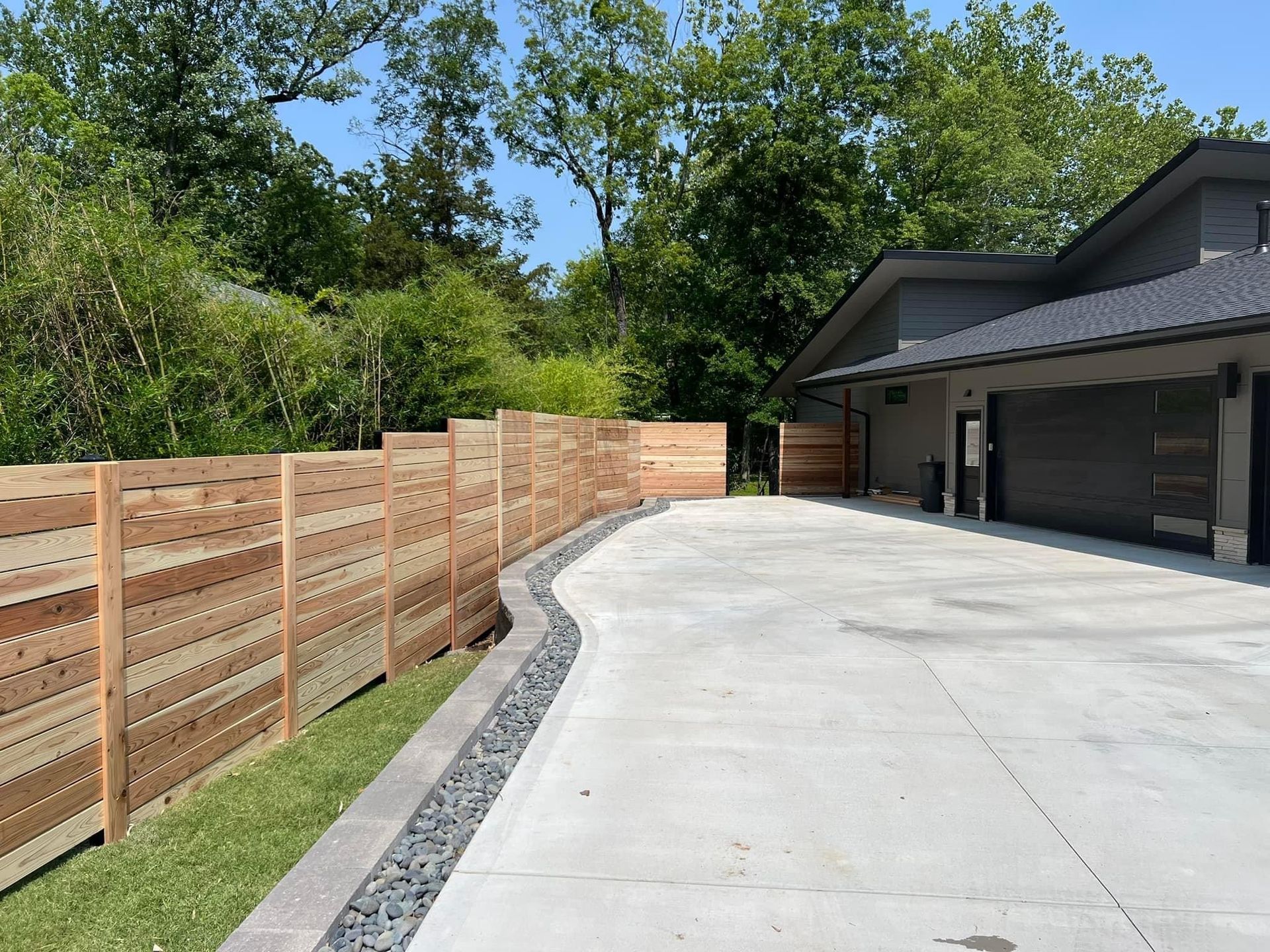 Curved concrete driveway beside a wood fence and house with garage, bordered by gravel and trees