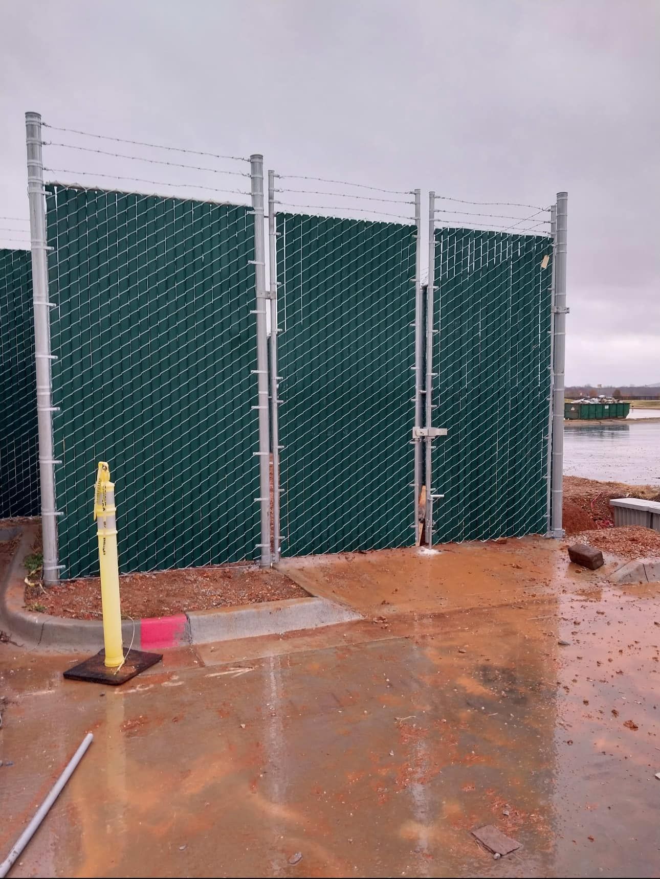 Green chain-link fence panels on a wet concrete pad in a rainy outdoor construction area.