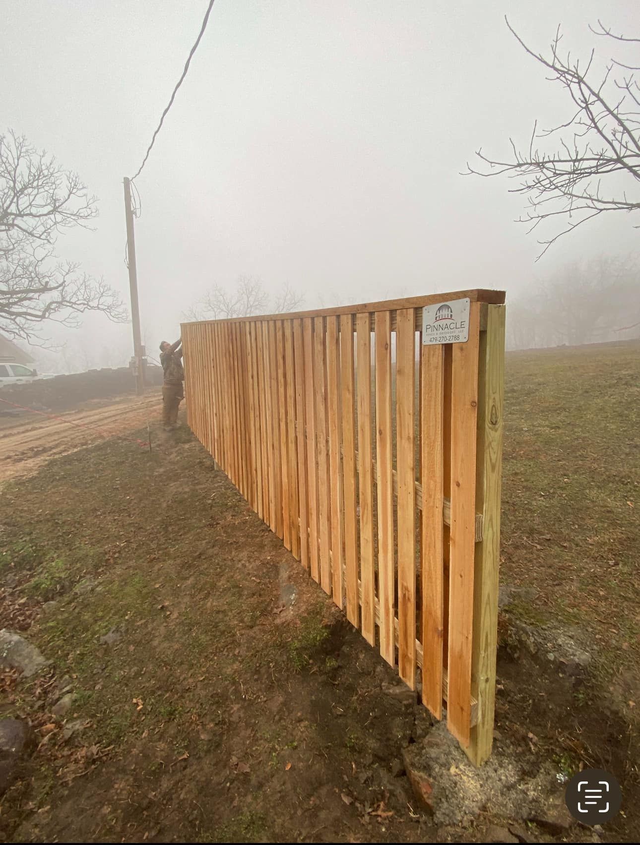 Foggy roadside scene with a long wooden fence and a utility pole beside a path.