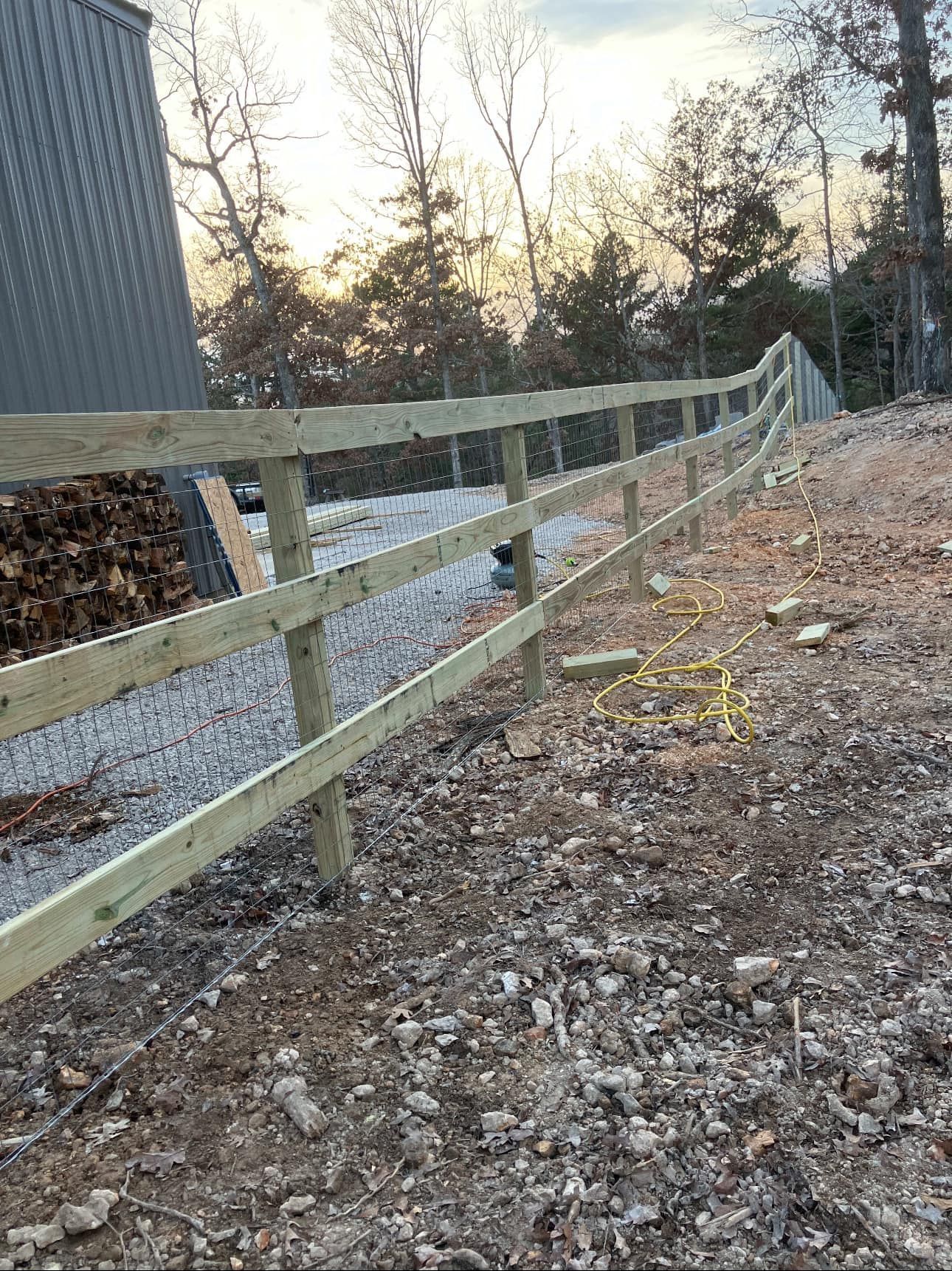 Wooden fence beside a gravel path near a building, with trees at sunset.