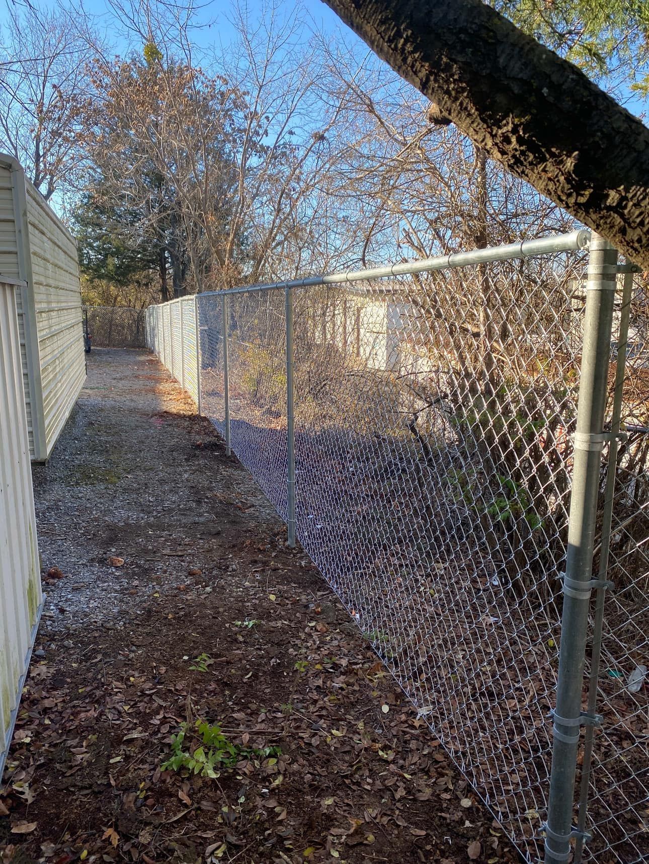 Narrow gravel path between a fence and chain-link fence, lined with fallen leaves under bare trees