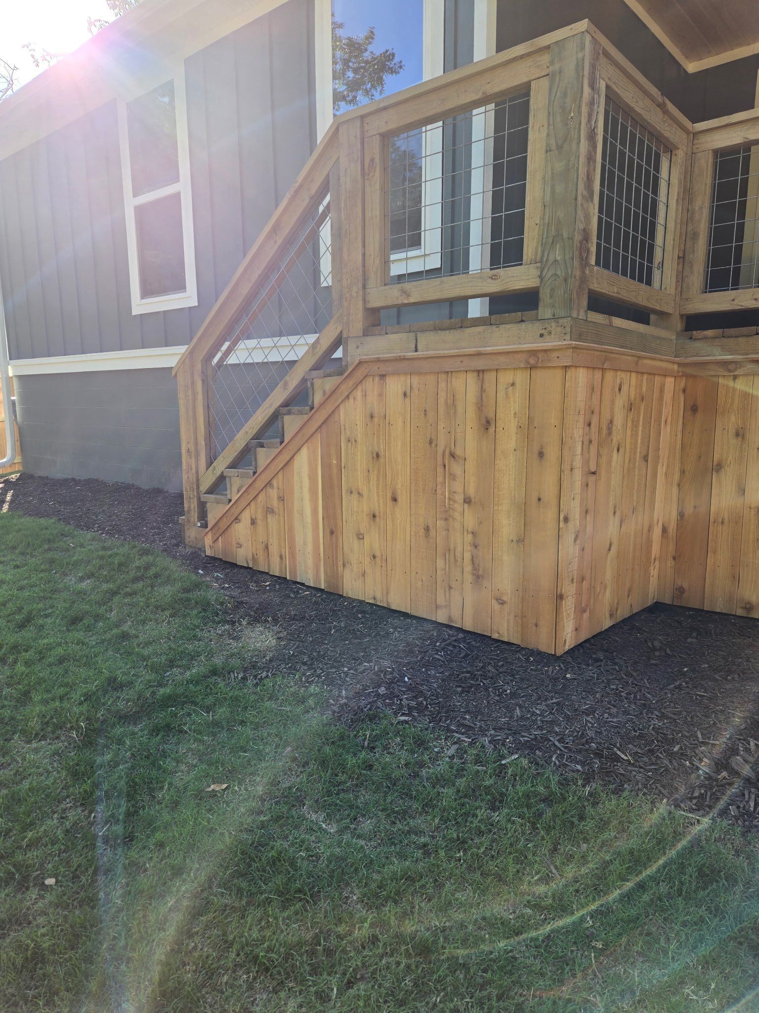 Wooden deck stairs and railing attached to a house, with sun glare and a grassy yard below.