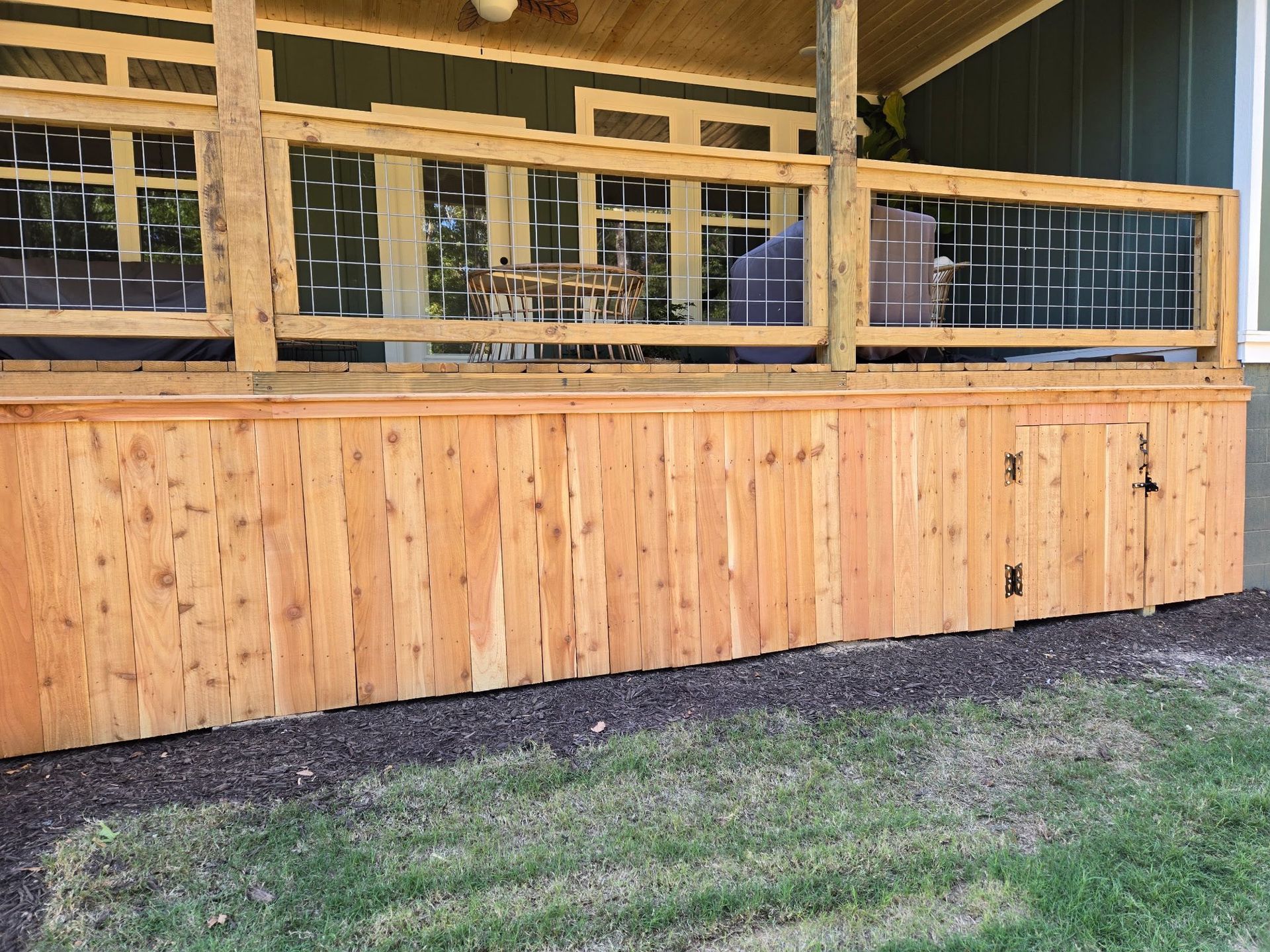 Wooden porch and deck with railing above a boarded lower wall beside a grassy yard
