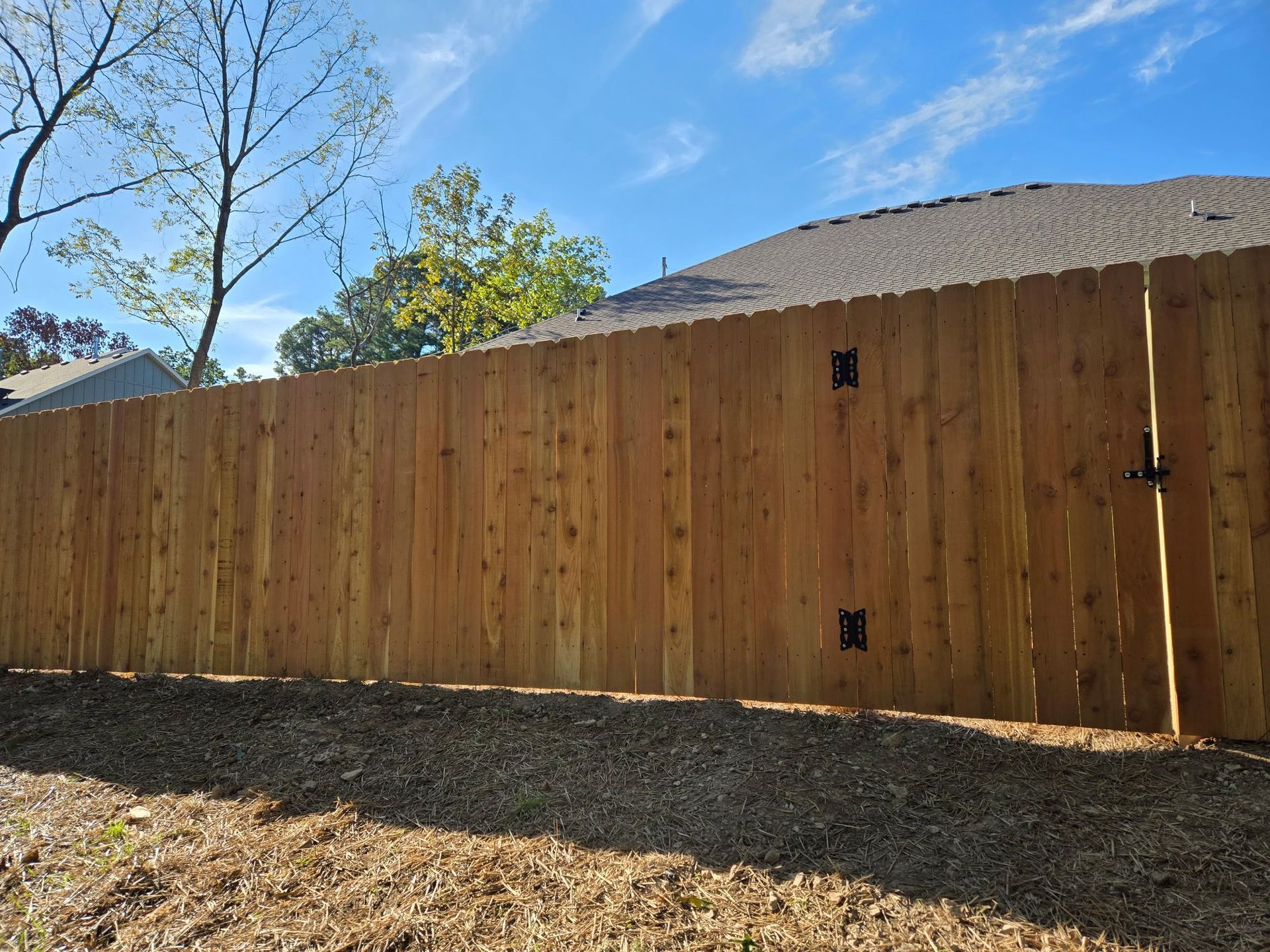 Wooden privacy fence with a gate in a sunny backyard, with trees and a house roof behind it