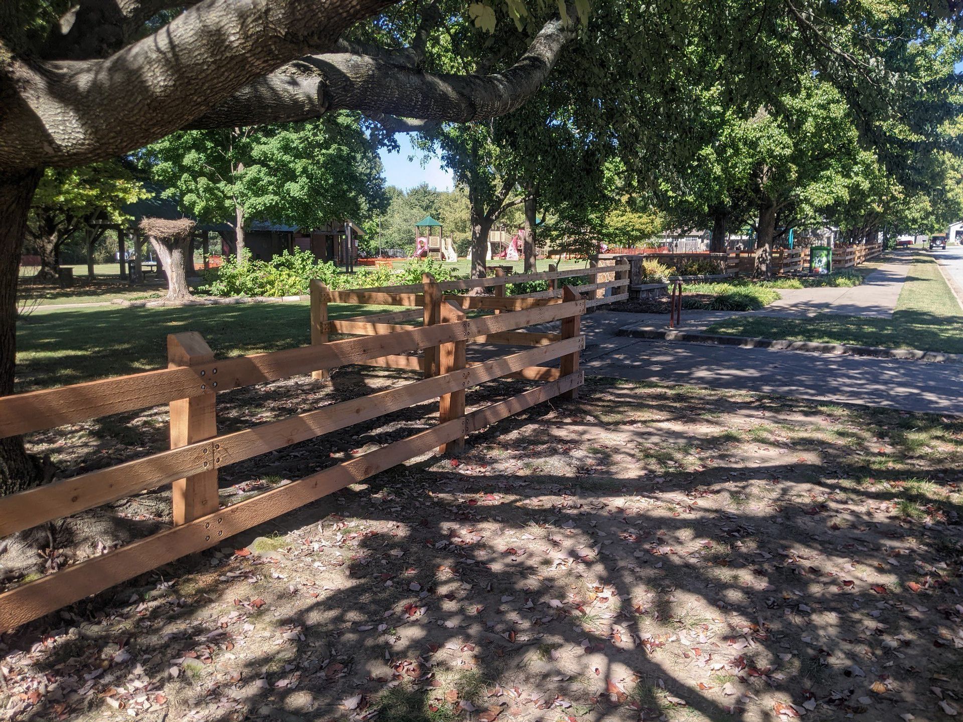 Wooden fence beside a shaded park path under a large tree, with dappled sunlight on the ground.