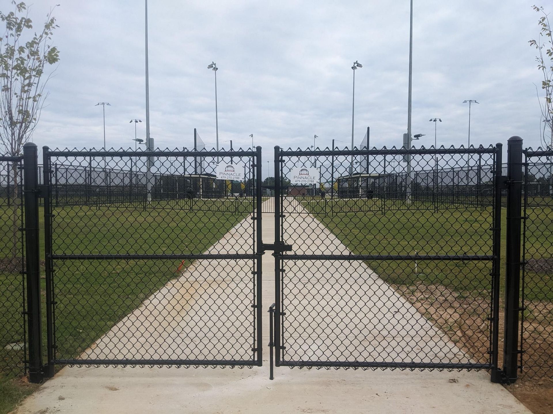 Chain-link gate opening to a concrete path in a fenced grassy park under a cloudy sky