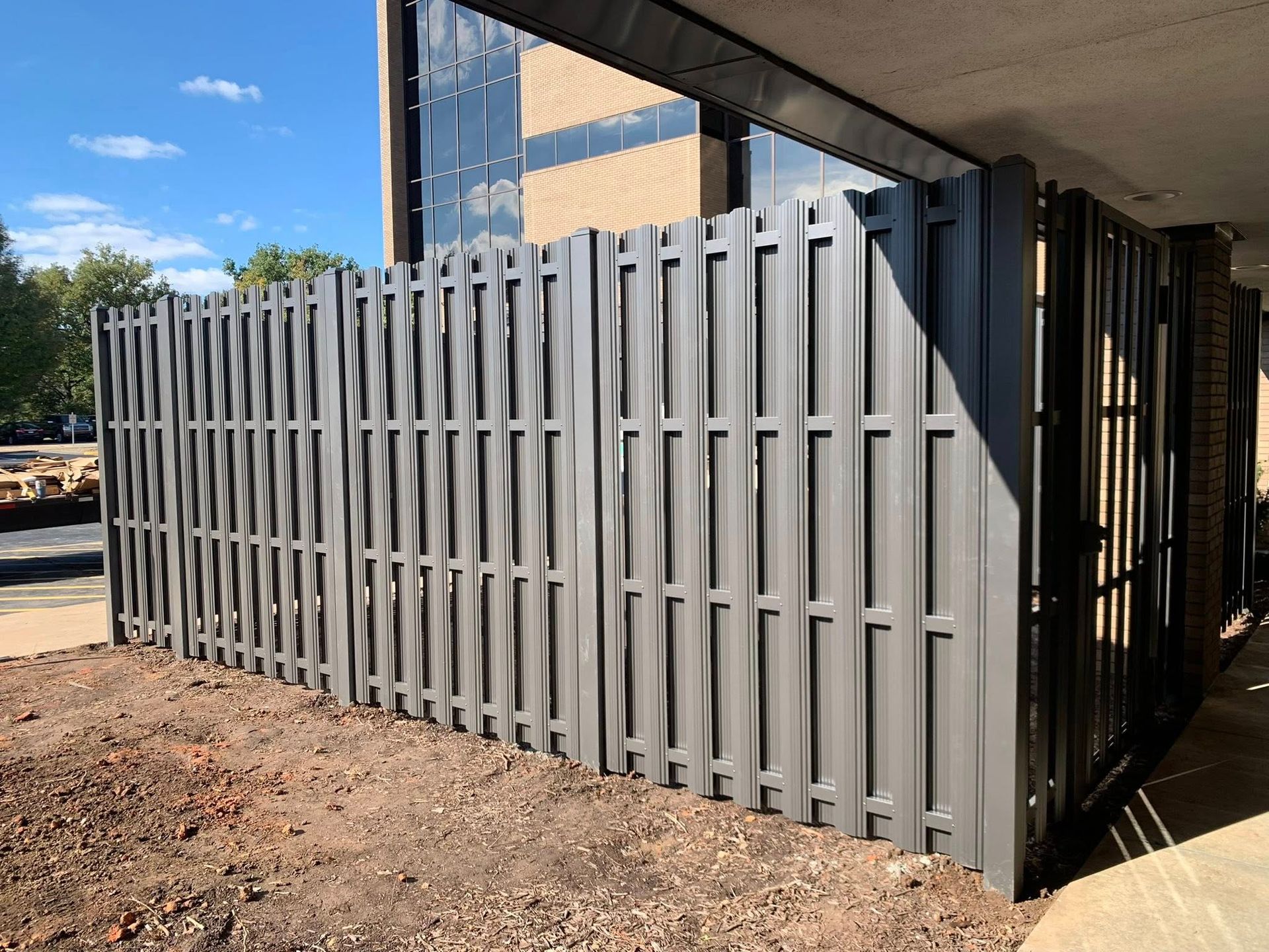 Gray metal slatted fence corner beside a building under construction, with dirt ground and blue sky.
