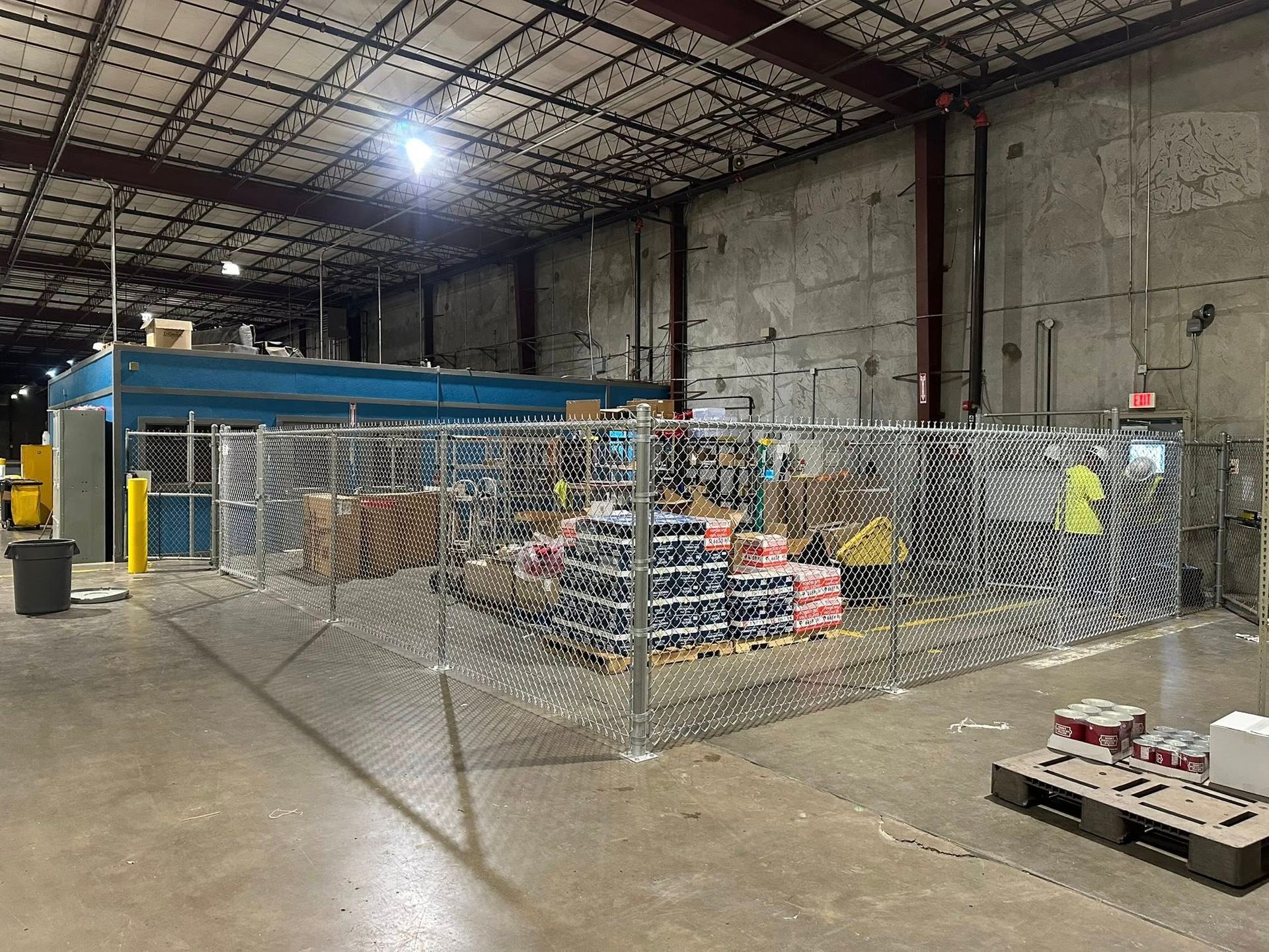 Warehouse interior with fenced storage area, blue machinery, pallets, and boxes under high ceiling lights