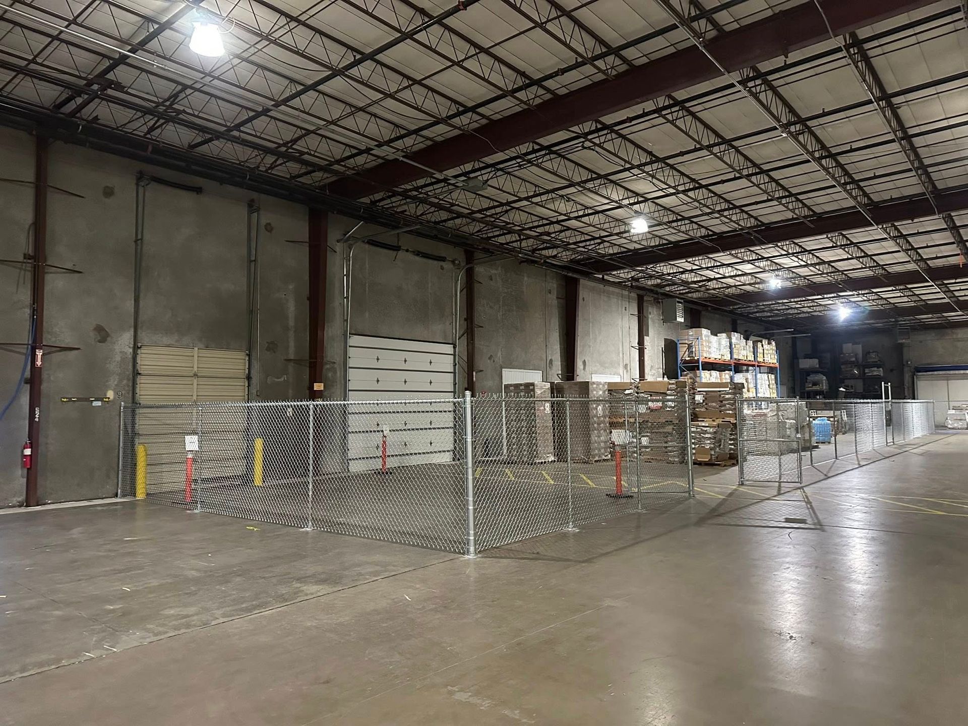 Empty warehouse interior with concrete floor, metal fencing, and stacked pallets under bright overhead lights