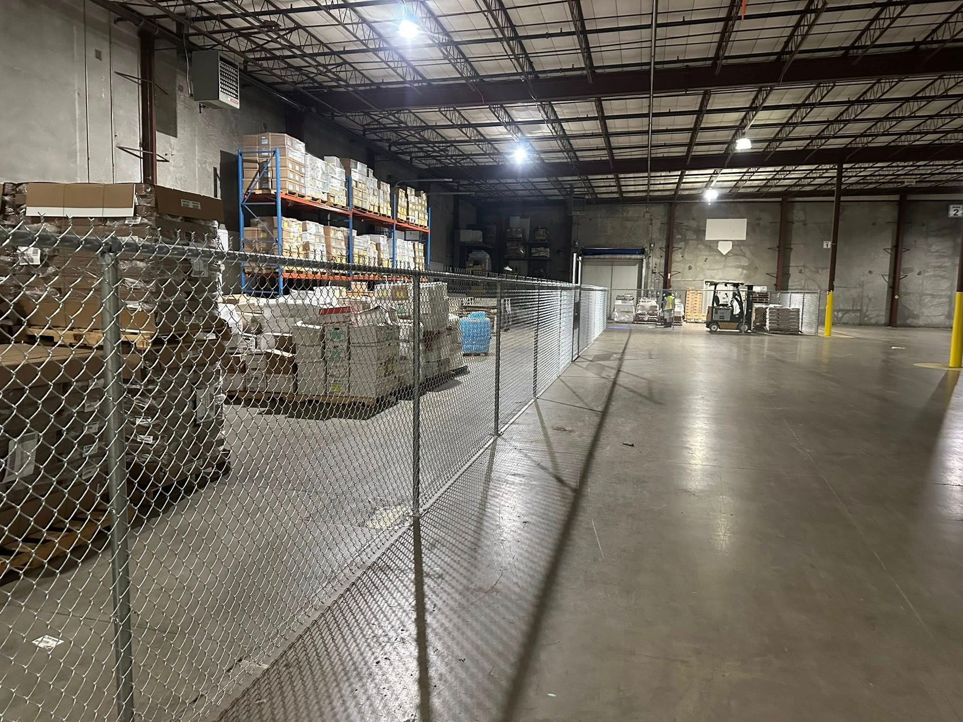 Industrial warehouse interior with chain-link fencing, stacked pallets, and overhead lights