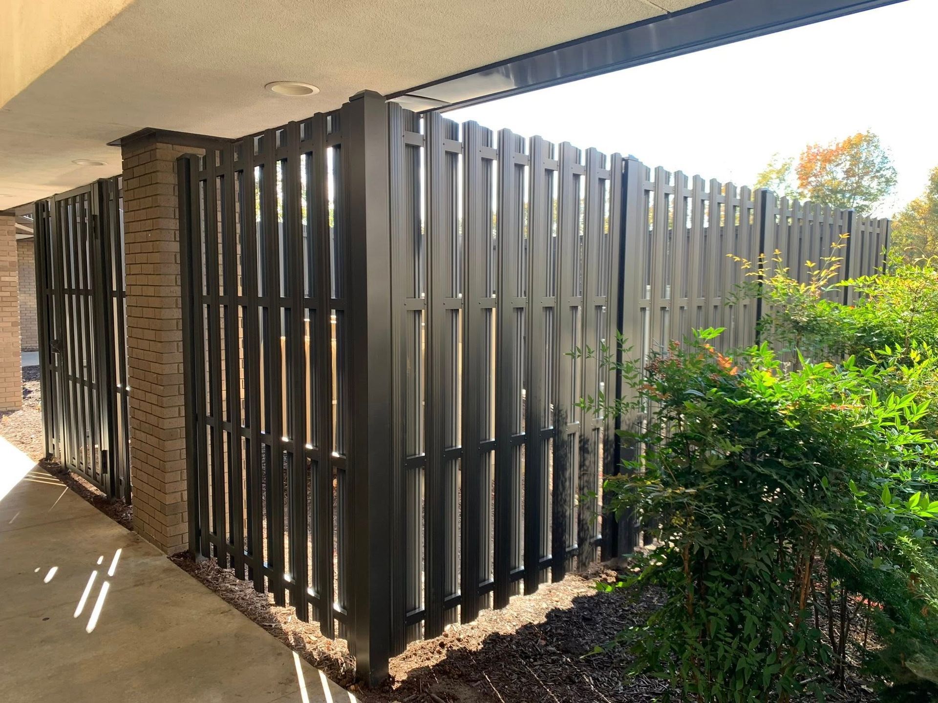 Dark slatted wooden fence beside a concrete walkway and green shrubs along a building exterior