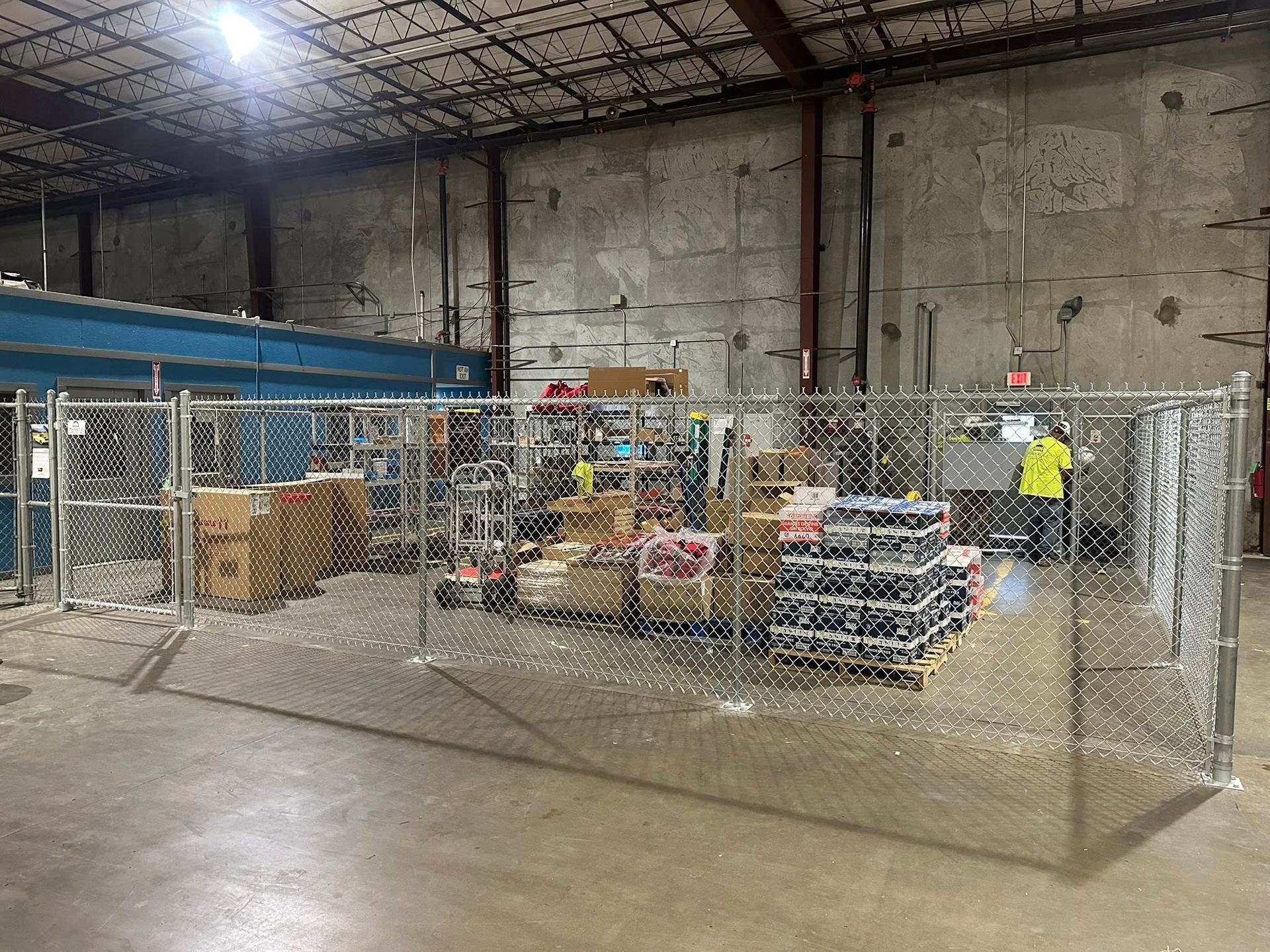 Warehouse storage area with chain-link pens, stacked boxes, and a worker in a yellow safety vest