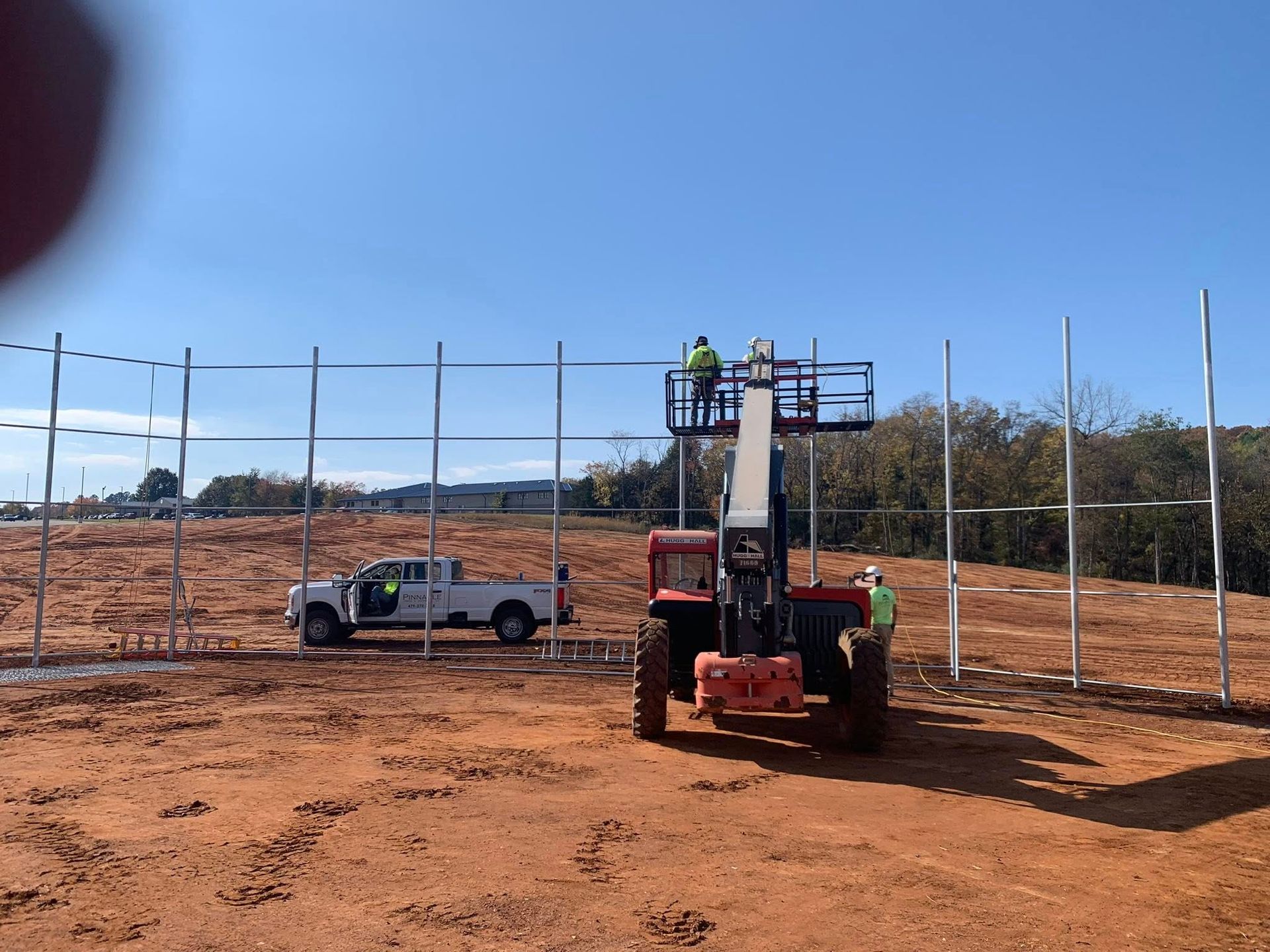 Construction site with a red lift and a white pickup beside metal fence posts under a blue sky