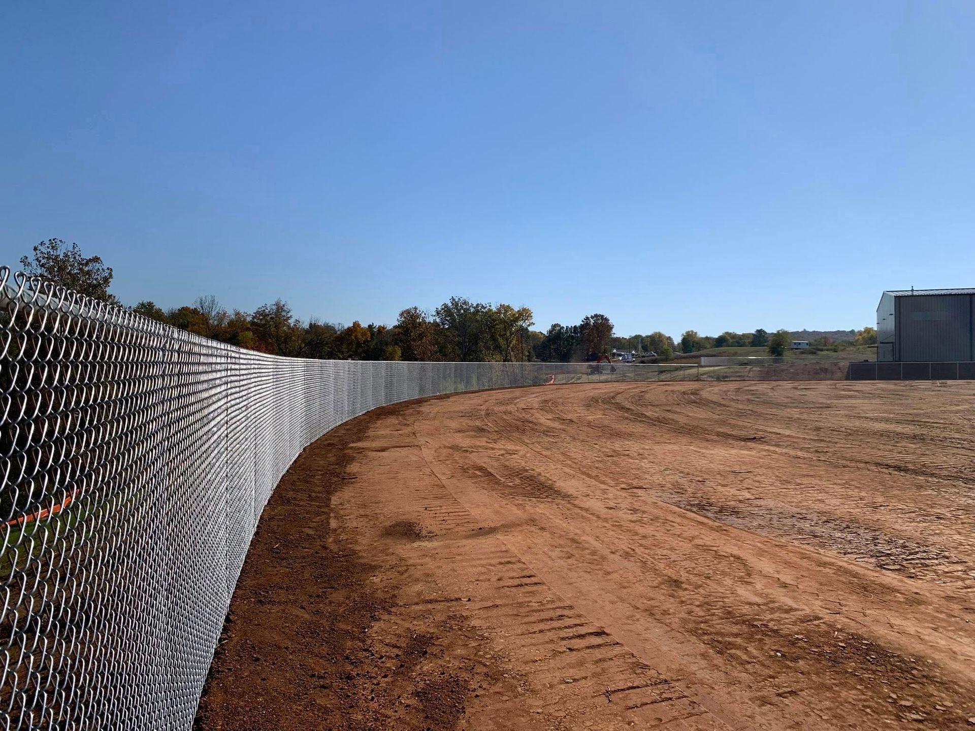 Chain-link fence along a dirt path beside an open field under a clear blue sky