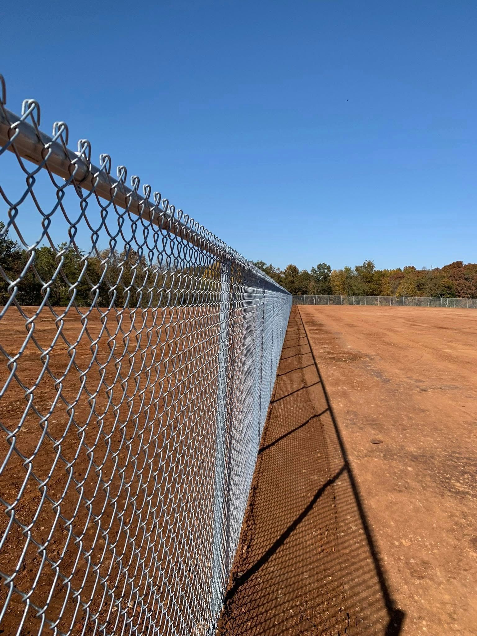 Chain-link fence along a dirt field under a clear blue sky, casting a long shadow.