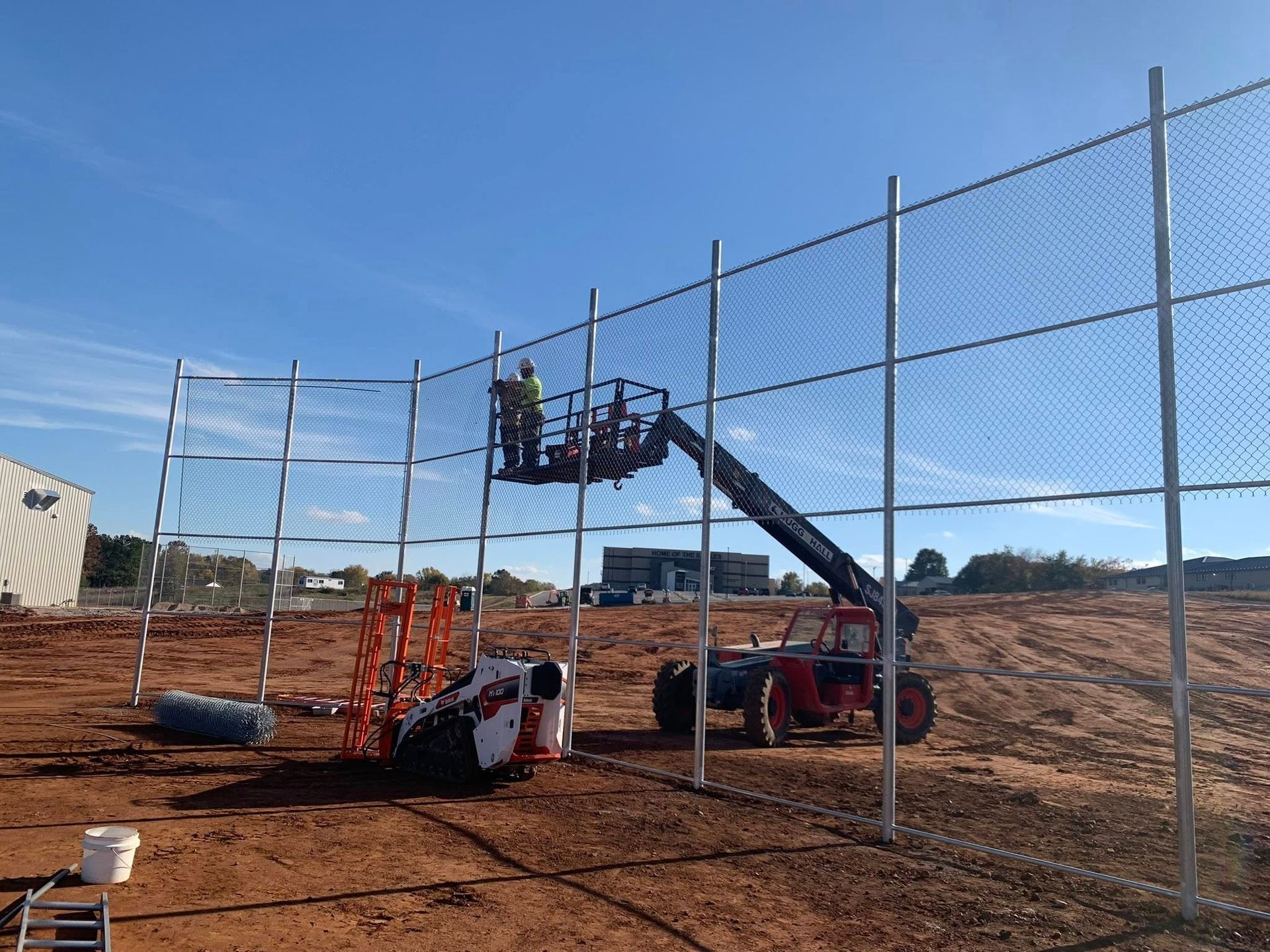 Workers on a lift installing netting on a tall sports fence at a construction site.