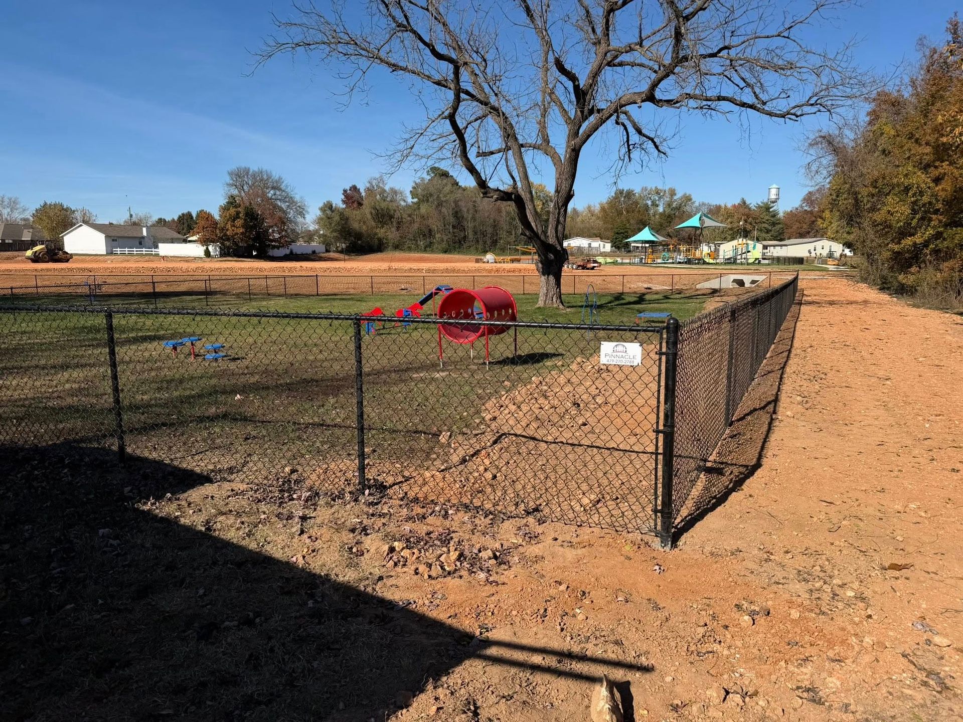 Fenced dirt enclosure with a red play tunnel on a grassy field under a leafless tree, on a sunny day