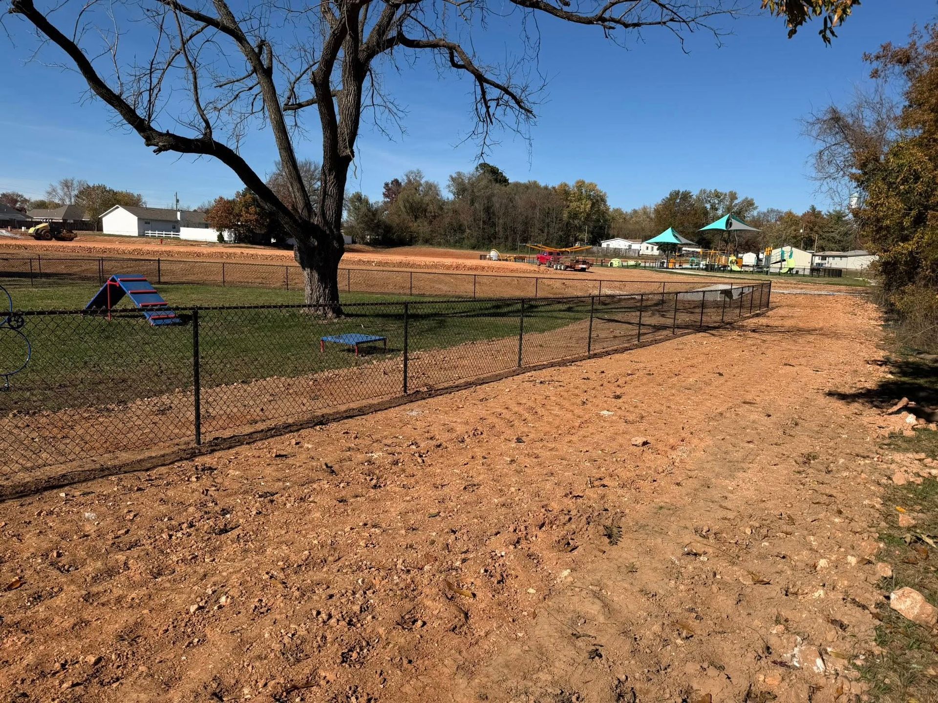 Fenced park with dry grass, bare trees, and small playground equipment under a clear blue sky