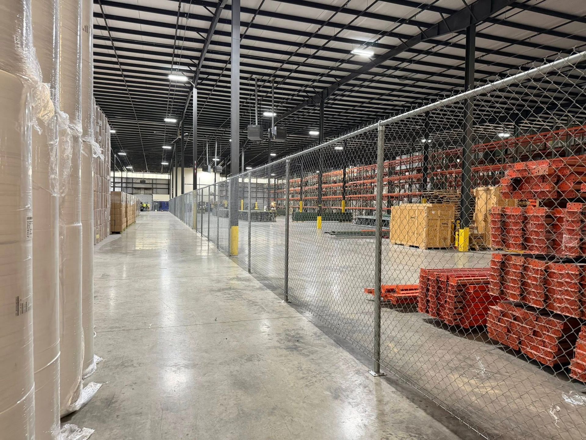 Warehouse aisle with chain-link fence, stacked red pallets, and tall shrink-wrapped goods