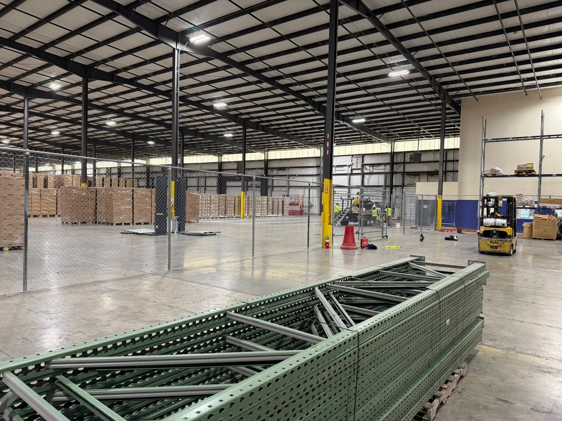 Large warehouse interior with metal trusses, stacked pallets, and a green steel frame in the foreground.