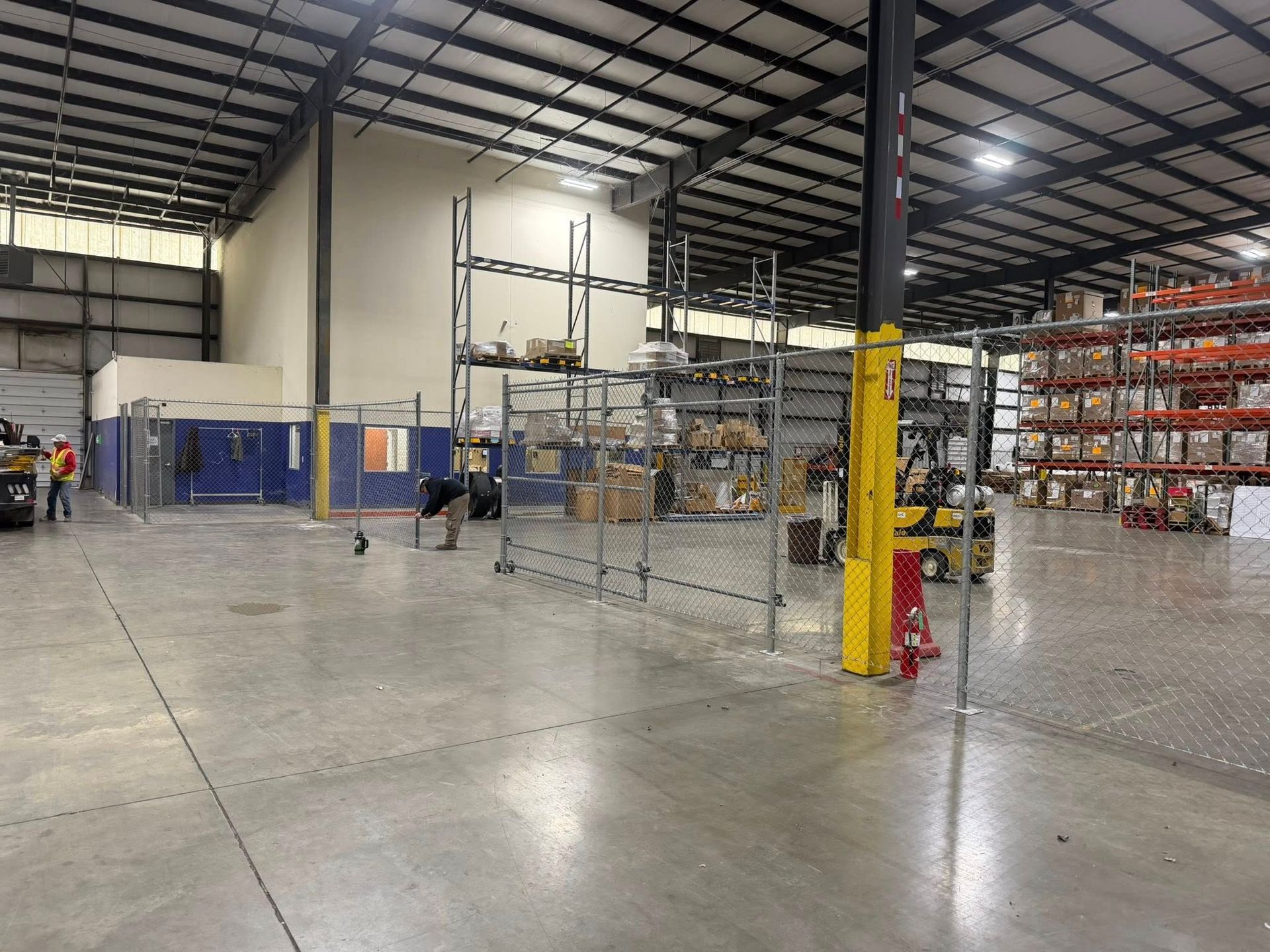 Large warehouse interior with fenced storage area, metal shelving, and two workers near the back wall