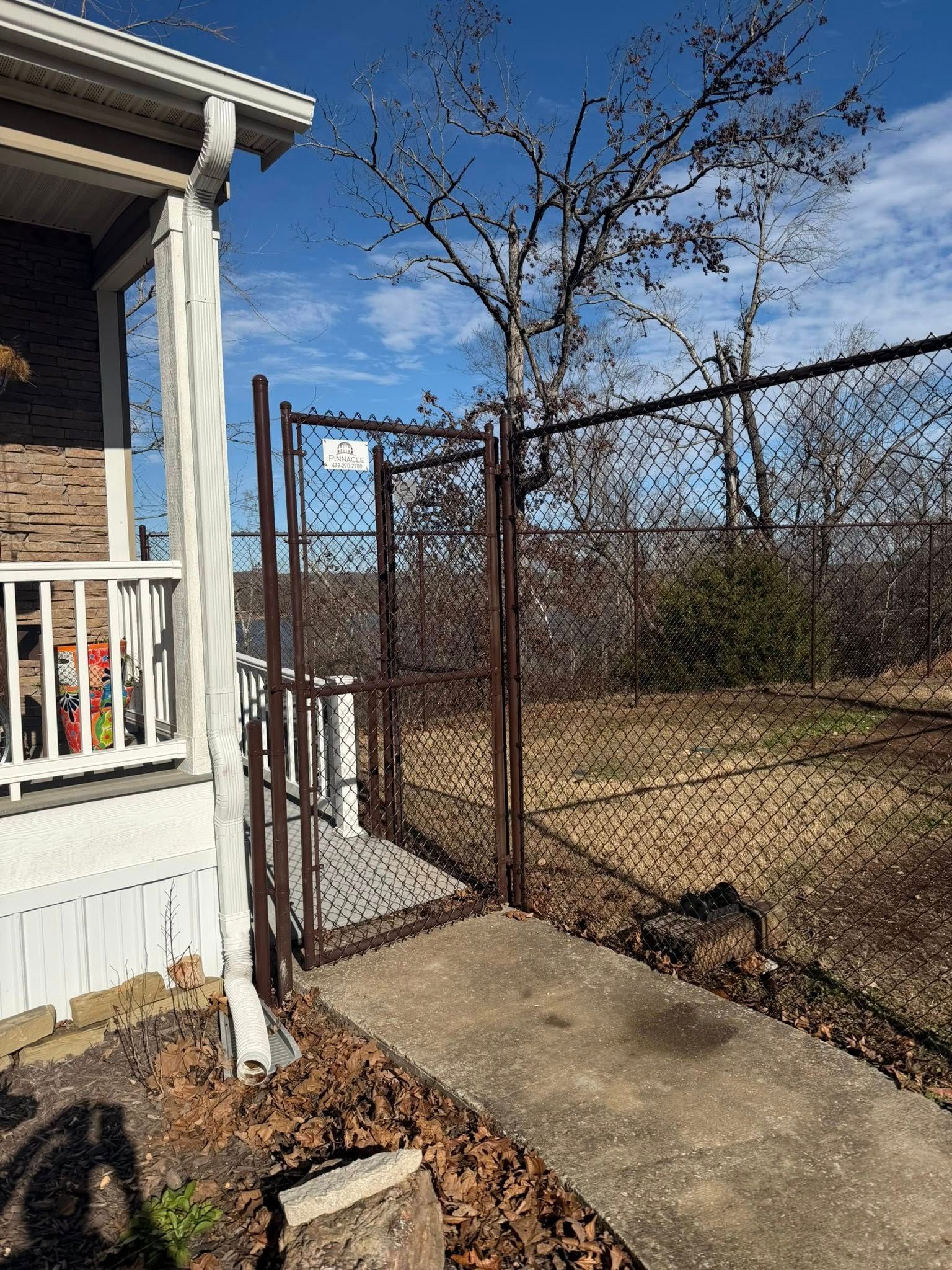 Narrow concrete walkway beside a white porch and chain-link fence, with dry yard and leafless tree under blue sky