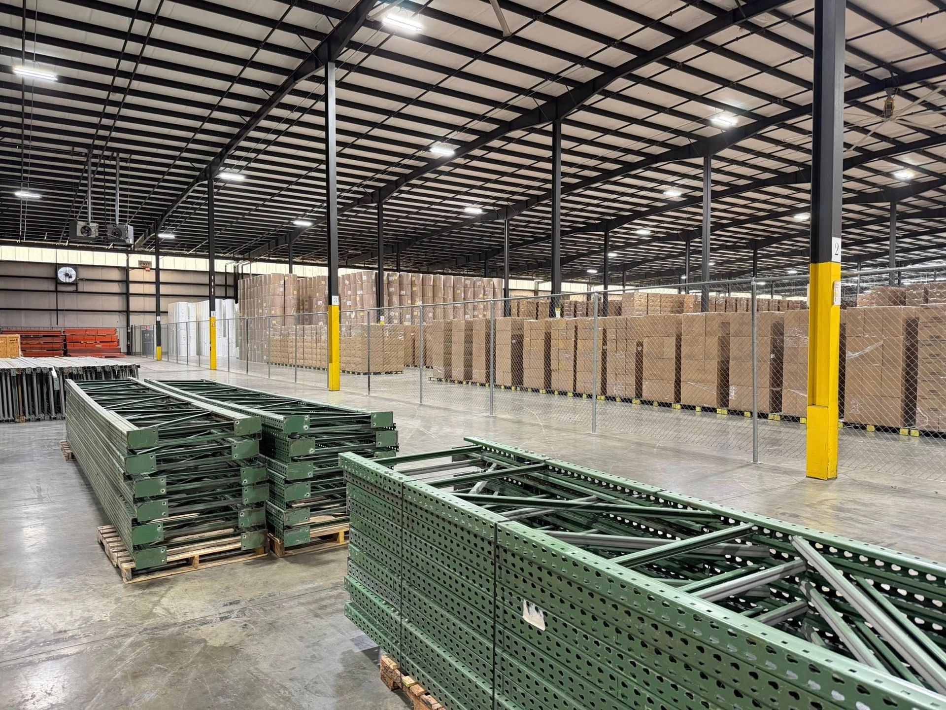 Warehouse interior with stacked pallets, green metal racks, and rows of boxed goods under a high ceiling