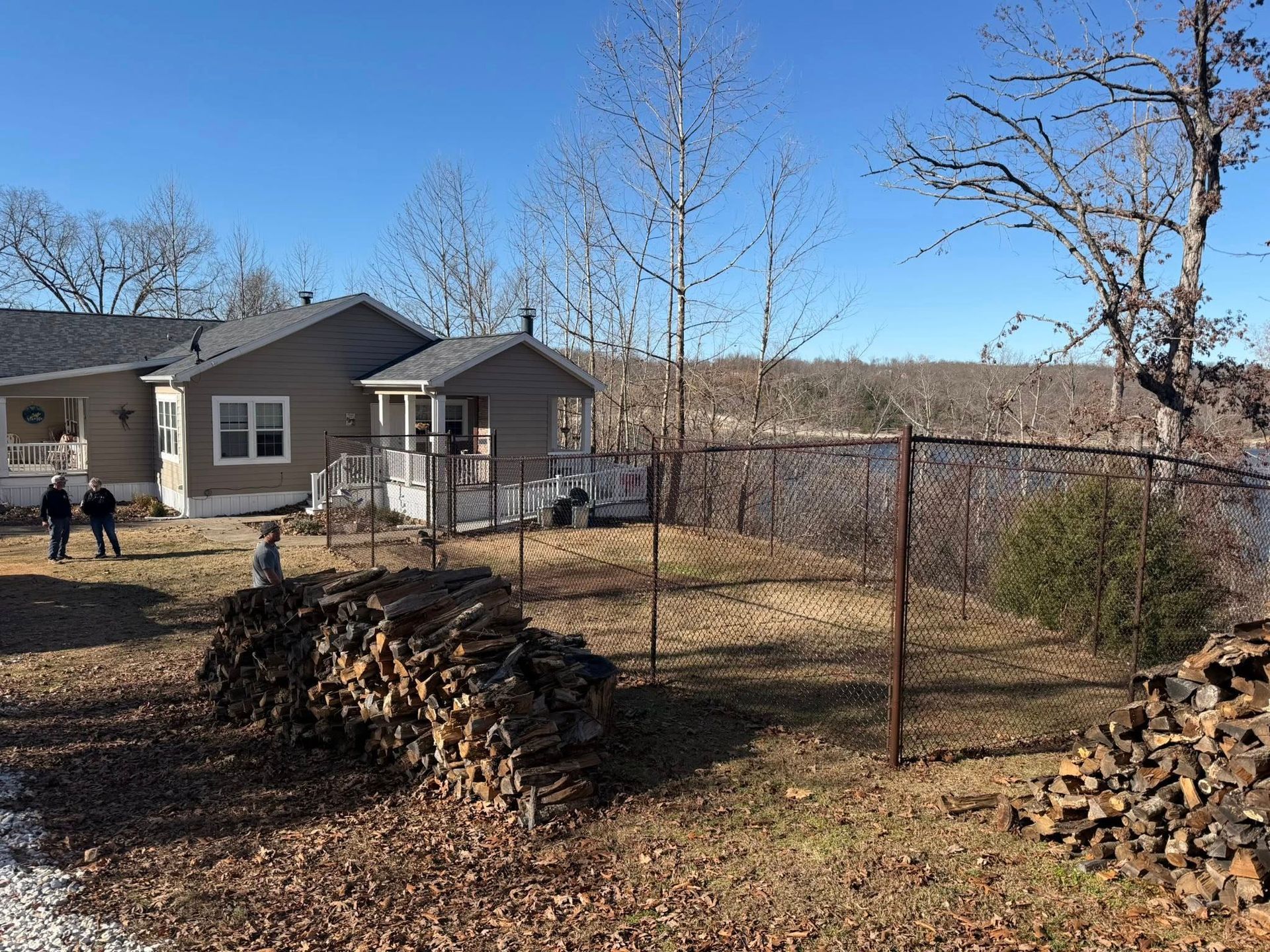 Backyard with stacked firewood, bare trees, a small house, and a wire fence under a clear blue sky