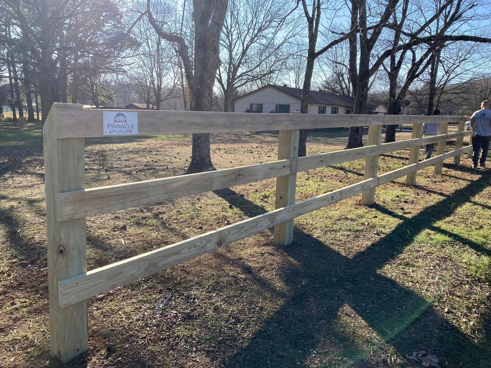 Wooden fence beside a grassy yard in winter, with a person walking in the distance and a house behind it