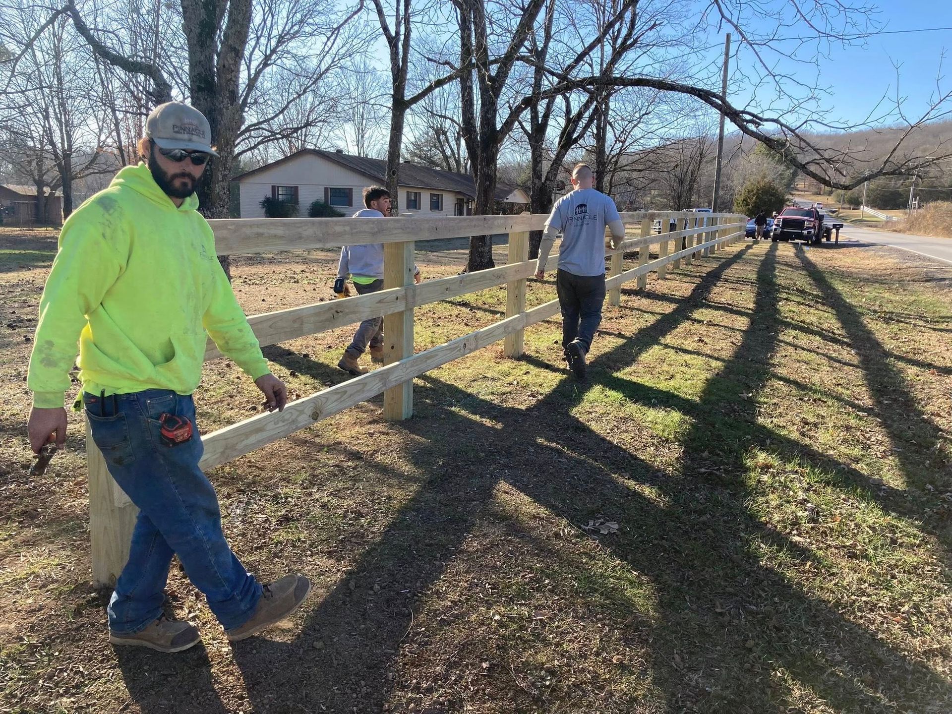 Three people painting a white fence along a grassy yard on a sunny day